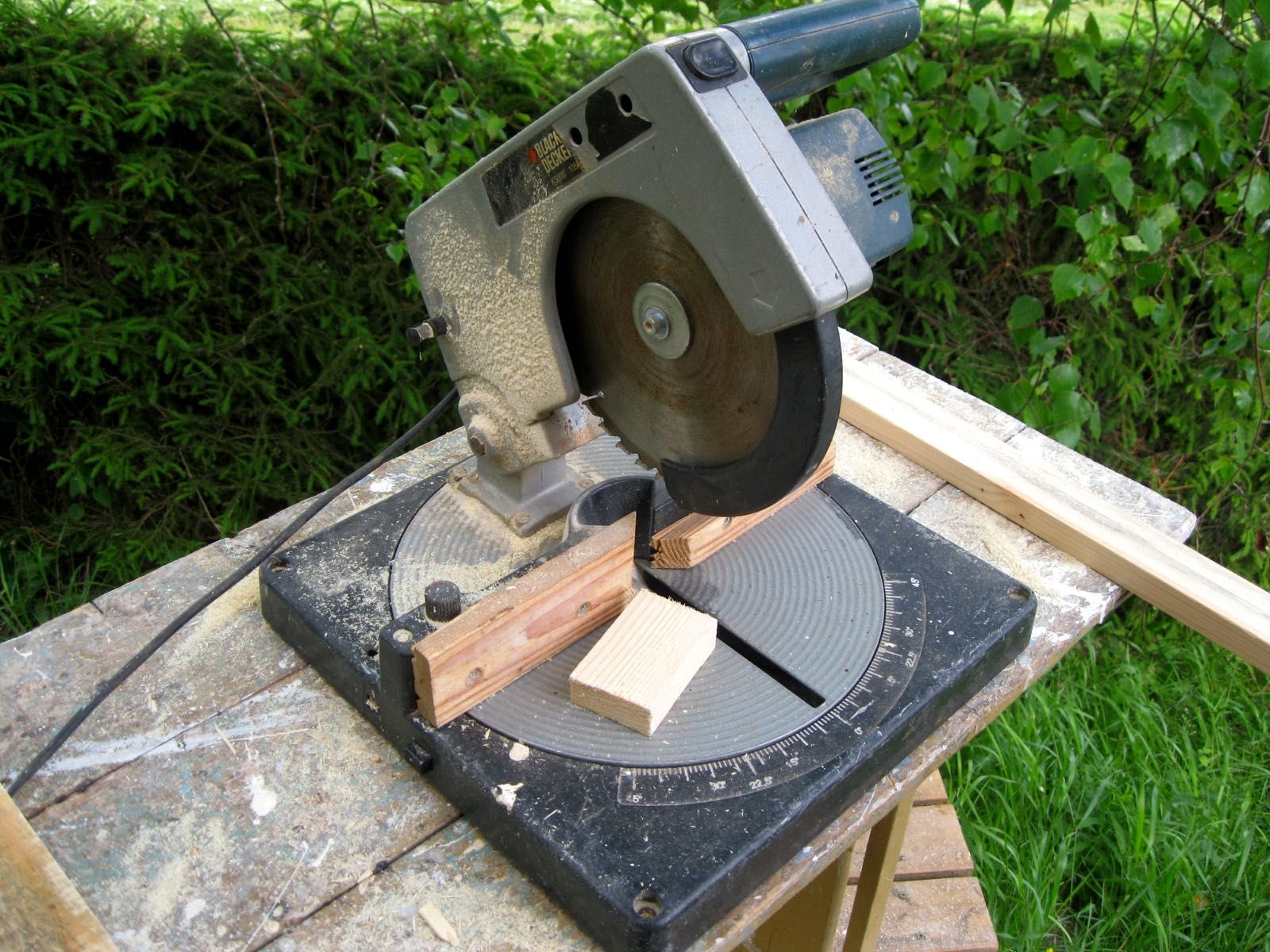 An older mitre saw on a garden workbench, with a wooden board screwed to the fence on both sides of the blade and an offcut block on the table