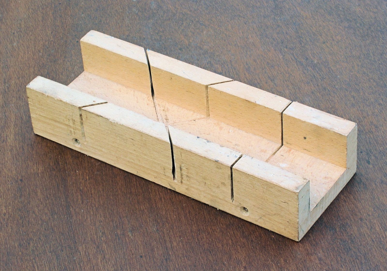 A wooden mitre box on a table: a bottom and two sides, with slots cut at 45 degrees both ways and at a right angle